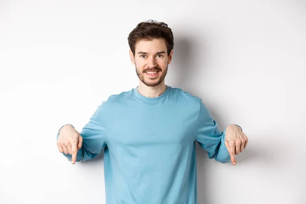 Sonriente hombre confiado apuntando con los dedos hacia abajo, mostrando banner de promoción o logotipo en el fondo blanco, de pie en sudadera azul casual — Foto de Stock