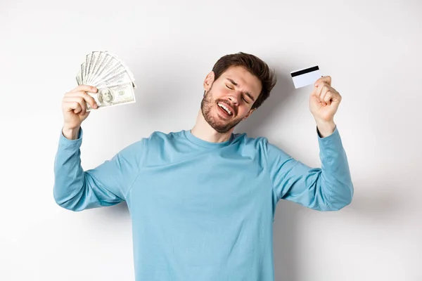 Hombre guapo bailando con dinero y tarjeta de crédito de plástico, de pie en ropa casual sobre fondo blanco — Foto de Stock