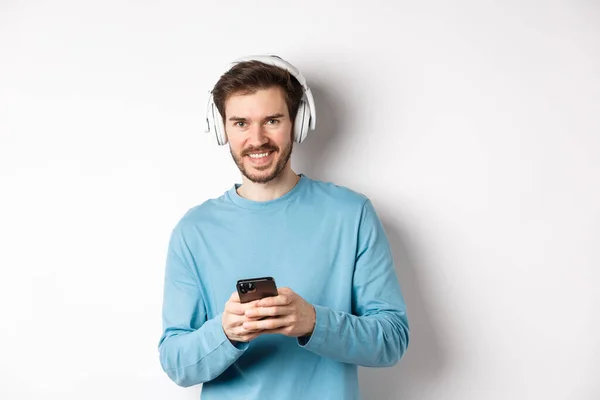 Homem sorridente atraente ouvindo música em fones de ouvido sem fio, usando smartphone preto e olhando satisfeito, fundo branco — Fotografia de Stock