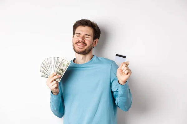 Chico alegre celebrando salario, bailando con tarjeta de crédito de plástico y dinero, sonriendo satisfecho, de pie sobre fondo blanco — Foto de Stock