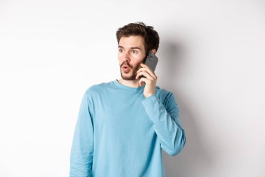 Teknoloji konsepti. İlgi çekici genç adam soldan etkilenmiş görünüyor, cep telefonuyla konuşuyor, birini arıyor, beyaz arka planda duruyor.