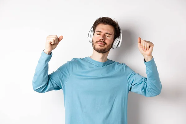 Homem bonito feliz dançando à música em fones de ouvido, ouvindo músicas e sorrindo, de pé sobre fundo branco — Fotografia de Stock
