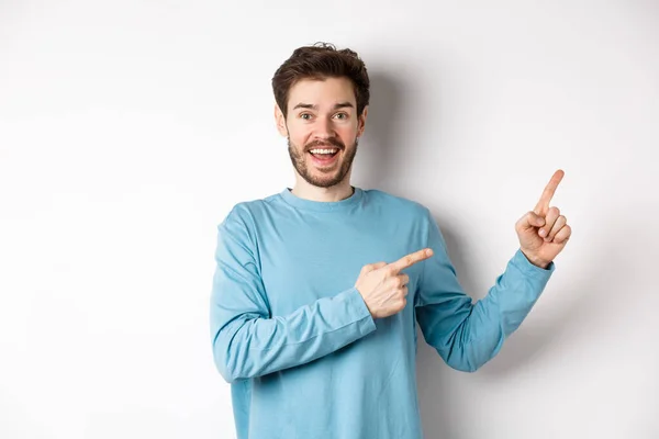 Веселый улыбающийся мужчина, указывающий пальцами направо и показывающий баннер или логотип на белом пространстве для рекламы, рекомендующий продукт, стоящий на фоне студии — стоковое фото