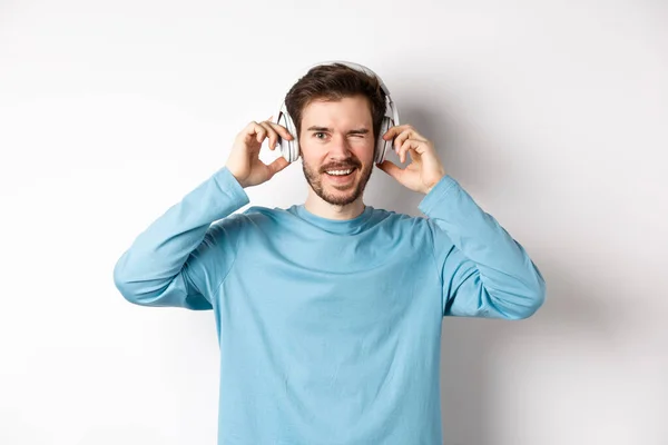 Glada kille blinkar åt kameran och ler, lyssna musik i trådlösa hörlurar, vit bakgrund — Stockfoto