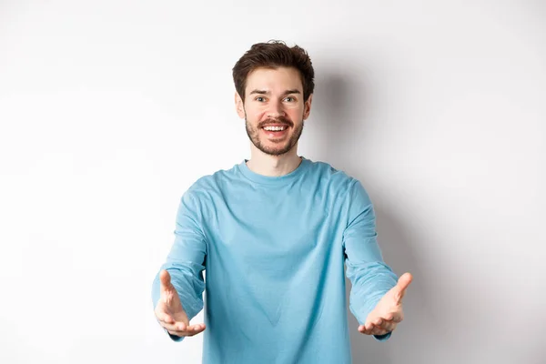 Привлекательный кавказский парень в повседневной одежде протягивает руки, чтобы держать что-то, принимая и улыбаясь, стоя на белом фоне — стоковое фото