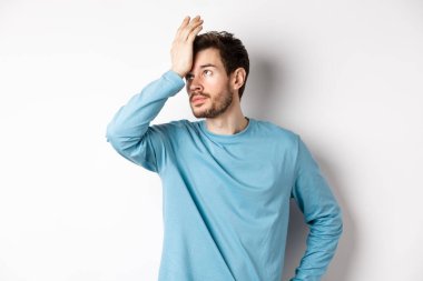 Annoyed young man with beard, roll eyes and making facepalm, pissed-off with something, standing irritated on white background