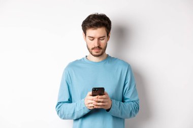 Akıllı telefondan mesaj okuyan, beyaz arka planda duran, mobil ekrana bakan ciddi bir genç adam.