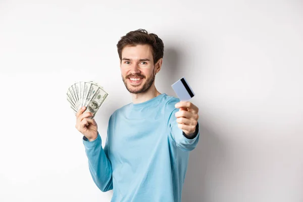Sonriente hombre caucásico sosteniendo el dinero y dándole tarjeta de crédito de plástico, de pie sobre fondo blanco — Foto de Stock