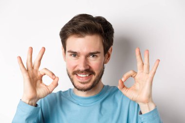 Kendine güvenen, gülümseyen, iyi işaretler gösteren, mükemmel seçimi öven, iyi bir şeyi onaylayan, beyaz arka planda duran genç adamın yakın çekimi.