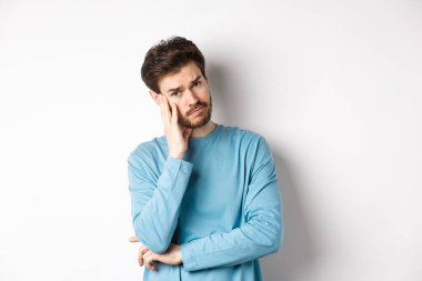 Üzgün genç adam sinirli görünüyor, baş tapınağına dokunuyor ve tedirgin, beyaz arka plana karşı duruyor.