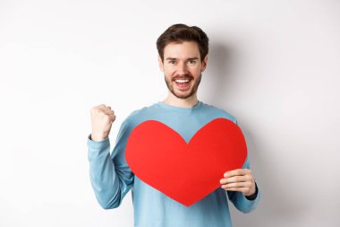 Sevgililer Günü 'nde aşkı bulmaya kararlı neşeli bir adam. Yumruk tokuşturuyor, başarılı jestler yapıyor ve beyaz arka planda duran büyük kırmızı romantik kalbi tutuyor.