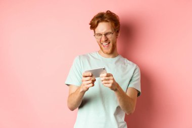Kırmızı dağınık saçlı, gözlüklü, akıllı telefondan video oyunu oynayan ve eğlenen, mobil ekrana bakan, pembe arka planda duran mutlu genç adam.