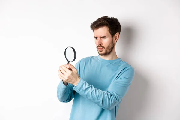 Serieus uitziende man kijkend door vergrootglas, iets onderzoekend, vond interessante promo, staand op witte achtergrond — Stockfoto