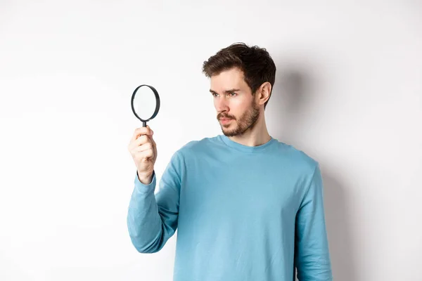 Young handsome man look through magnifying glass with curious face, investigating or searching for something, standing on white background — Stock Photo, Image