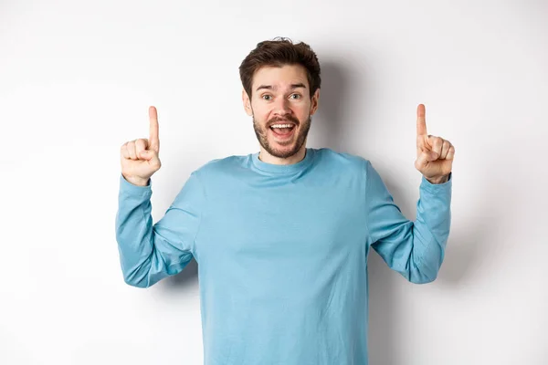 Веселый молодой человек, показывающий рекламу со счастливой улыбкой, указывающий пальцами на удивительный логотип баннера, стоящий на белом фоне — стоковое фото