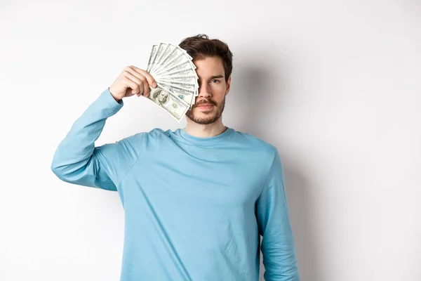 Joven serio cubriendo la mitad de la cara con dinero, mirando a la cámara, de pie en sudadera azul sobre fondo blanco — Foto de Stock