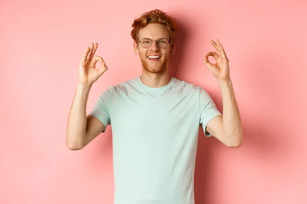 Hombre caucásico satisfecho con el pelo rojo, con gafas y camiseta, mostrando gestos bien y sonriendo, diciendo sí y aprobando algo, de pie sobre fondo rosa — Foto de Stock