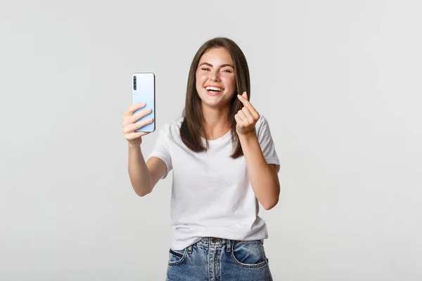 Onnellinen kaunis nuori nainen osoittaa sydämen elettä ja ottaa selfie älypuhelimella, nauraa huoleton — kuvapankkivalokuva
