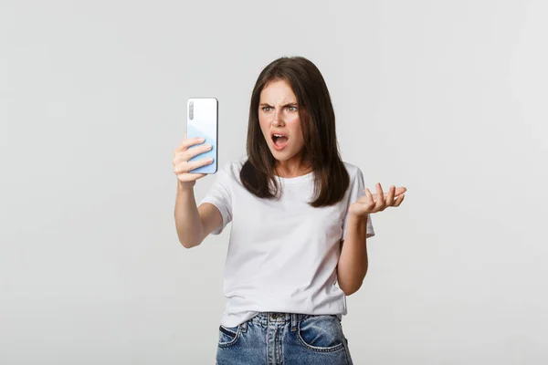 沮丧的年轻女人在视频电话上争吵，拿着智能手机，站在白色背景上 — 图库照片