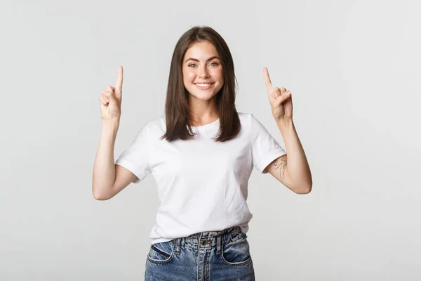 Menina morena sorridente atraente apontando os dedos para cima, mostrando logotipo — Fotografia de Stock