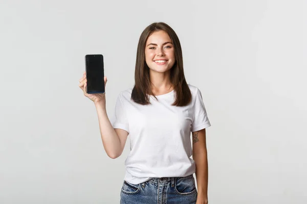 Attraktiv lächelnde junge Frau sieht zufrieden aus und zeigt Smartphone-Bildschirm — Stockfoto