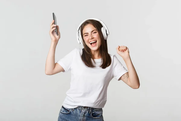Joyeux belle fille brune dansant et écoutant de la musique dans des écouteurs sans fil, tenant smartphone — Photo