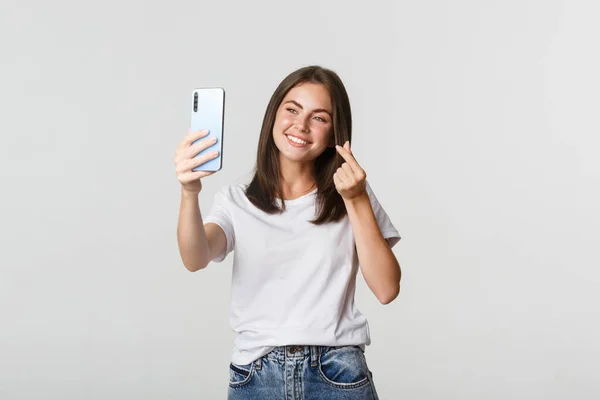 Glücklich schöne junge Frau zeigt Herz-Geste und macht Selfie auf Smartphone und lächelt — Stockfoto