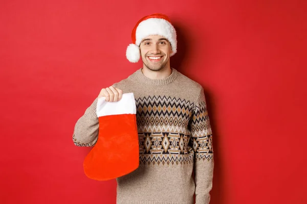 Koncept zimních prázdnin, Nový rok a oslavy. Obrázek pohledného usmívajícího se muže v Santa klobouku a svetru, držící vánoční punčochu na dárky a bonbóny, stojící nad červeným pozadím — Stock fotografie