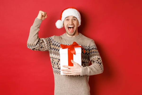 Bild eines glücklichen und aufgeregten gutaussehenden Mannes, der sich ein Weihnachtsgeschenk macht, triumphierend die Hände hebt und lächelnd das neue Jahr feiert, vor rotem Hintergrund in Weihnachtsmütze steht — Stockfoto