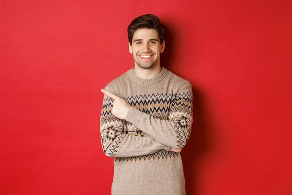 Bilde av kjekk mann i julegenser, feiring av nyttårsferier, smilende glad og pekefinger i øverste venstre hjørne kopieringsrom, stående over rød bakgrunn – stockfoto