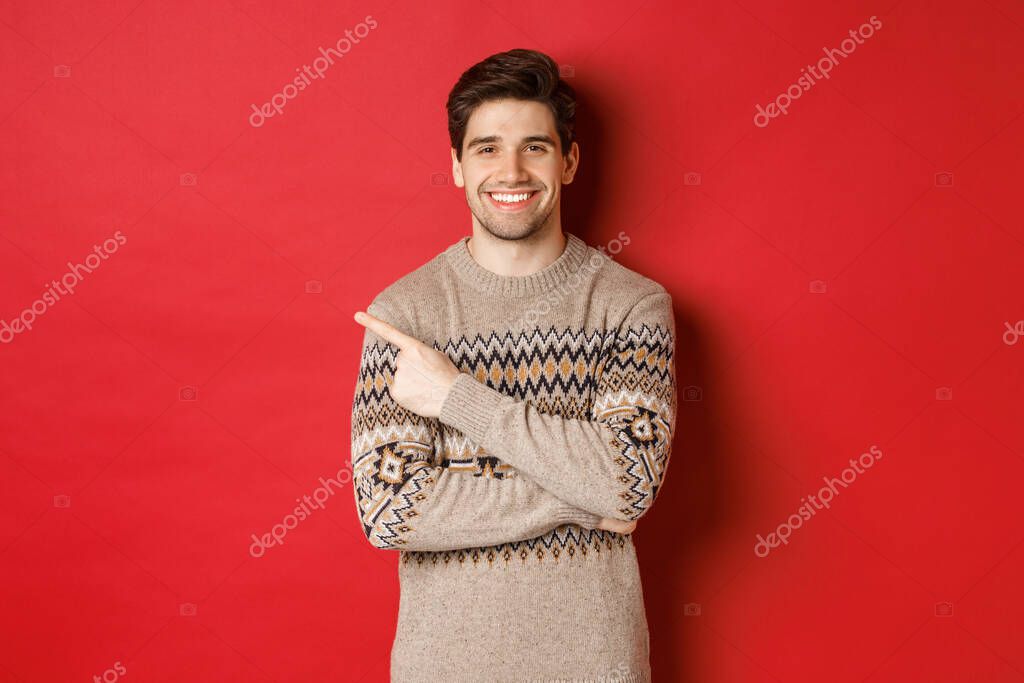 Image of handsome man in christmas sweater, celebrating new year holidays, smiling happy and pointing finger at upper left corner copy space, standing over red background.