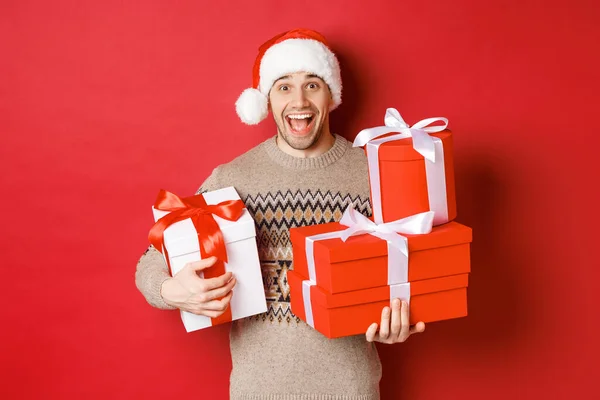 Konzept der Winterferien, Neujahr und Feier. Bild eines glücklichen Kerls, der Weihnachten genießt, Geschenke in der Hand hält und amüsiert lächelt, vor rotem Hintergrund steht — Stockfoto