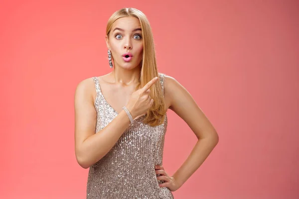 Wondered amused attractive blond woman talking party widen eyes say wow pointing questioned right direction see famous person standing surprised impressed gaze camera, red background