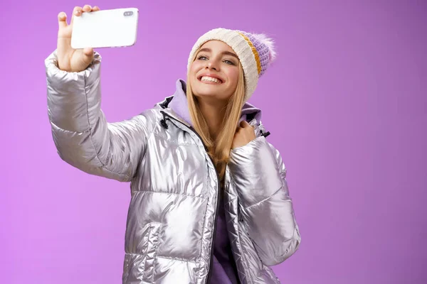 Glücklich entzückt unbeschwerte blondhaarige charmante Europäerin mit silberner Winterjacke, die ihr Smartphone waagerecht in die Höhe reckt und ein Selfie mit lächelndem Handydisplay macht, lila Hintergrund — Stockfoto