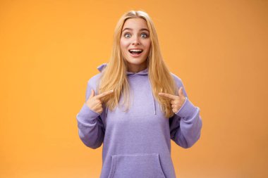 Şaşırdım genç sarışın kız kaşlarını kaldırdı kendini göstererek seçildi harika bir etkinliğe katıldım mutlu turuncu arka planda ayakta durarak şansa inanamıyorum