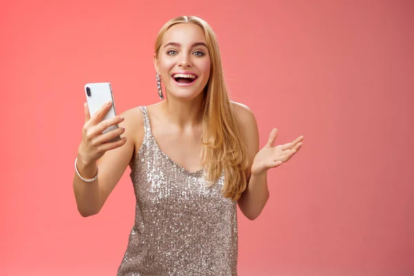 惊喜交集的金发美女穿着银光闪闪的时髦衣服，手持智能手机惊奇地喜欢令人敬畏的结果编辑照片应用程序笑得很开心，站在红色的背景上 — 图库照片