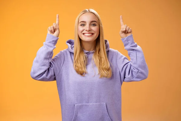 Φιλική χαρισματική energized νεαρή ξανθιά Ευρωπαίος γυναίκα σε ζεστό hoodie σηκώστε τα χέρια δείχνοντας τα δάχτυλα δείκτη παρουσιάζοντας φοβερό διαφήμιση χαμογελώντας ευτυχώς συνιστούμε προϊόν, πορτοκαλί φόντο — Φωτογραφία Αρχείου