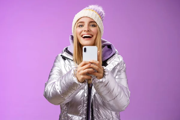 Amüsiert aufgeregt attraktive blonde Freundin hält Smartphone hoch Aufnahme Video Freund Schritt Snowboard zum ersten Mal Erfassung von Erinnerungen mobile Kamera steht glücklich lila Hintergrund — Stockfoto