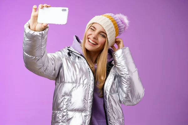Fröhlich unbeschwert charmante blonde Mädchen Spaß haben wollen Bilder aus dem Skiurlaub unter seflie halten Smartphone imitieren Show Zunge zwinkernden Blick mobiles Display, lila Hintergrund — Stockfoto