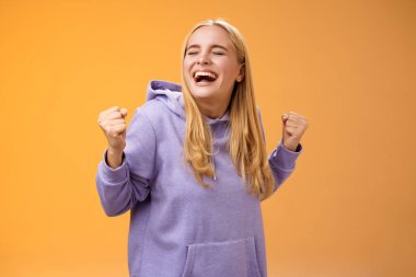 Şirin, mutlu, Avrupalı, kapüşonlu, sarışın, güzel bir kız, evet diye bağırıyor. Hayallerini gerçekleştirmek için yumruk sallıyor, zaferi kutluyor, amacına ulaşıyor, turuncu arka planda duruyor.
