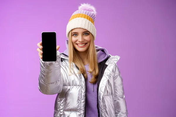 Freundlich fröhlich selbstbewusst blondes Mädchen in silberner stylischer Winterjacke Hut ausstrecken Arm zeigt Smartphone-Display Werbung awesome neue Gerät App lächeln selbstbewusst empfehlen Handy verwenden — Stockfoto