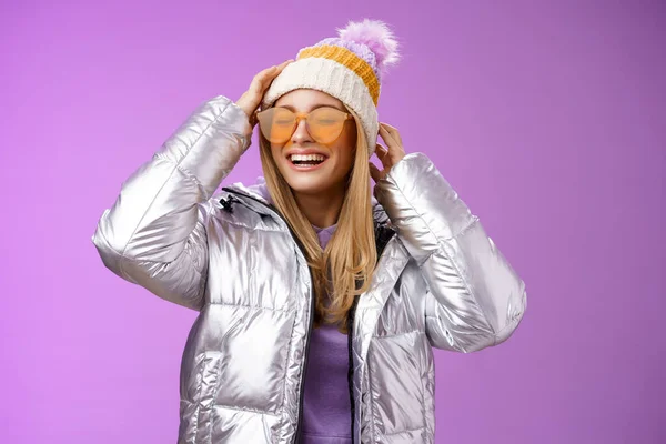 Despreocupado divertida novia rubia encantadora que se divierte disfrutando de impresionantes vacaciones soleadas vacaciones de invierno estación de esquí con gafas de sol chaqueta elegante de plata poner en el sombrero sonriendo alegremente, fondo púrpura —  Fotos de Stock