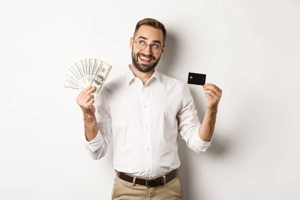 Imagen de un joven empresario con tarjeta de crédito y dinero, mirando a la esquina superior izquierda y pensando en ir de compras, de pie sobre fondo blanco — Foto de Stock