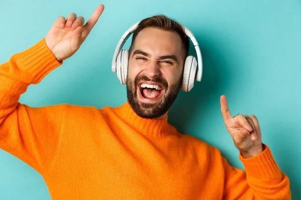Close-up de belo homem moderno ouvindo música em fones de ouvido, de pé em suéter laranja sobre fundo turquesa — Fotografia de Stock
