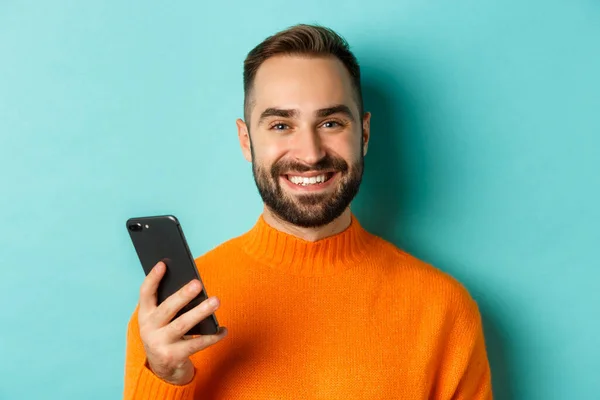 Primer plano del hombre guapo feliz escribiendo un mensaje en el teléfono móvil, sosteniendo el teléfono inteligente y sonriendo, de pie sobre fondo turquesa —  Fotos de Stock