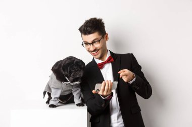 Yakışıklı genç adam köpeğine cep telefonundan bir şey gösteriyor. Sahibi evcil hayvanla online alışveriş yapıyor, beyaz arka planda kostümler içinde duruyor.