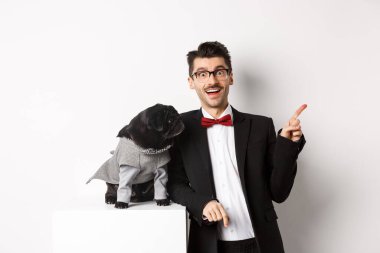 Hayvanlar, parti ve kutlama konsepti. Mutlu ve şaşkın genç adam ve kostümlü sevimli köpek sahibi beyaz arka planda dikiliyor.