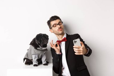 Takım elbiseli neşeli köpek sahibi köpekle yeni yılı kutluyor. Akıllı telefondan özçekim yapıyor. Kostümlü, beyaz arka planlı, sevimli siyah bir köpeğin yanında.