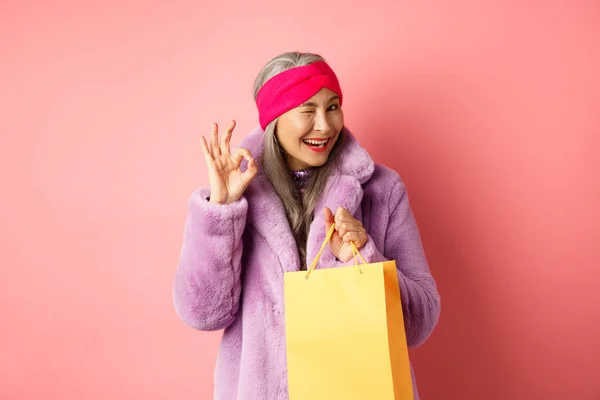 Stilvolle ältere asiatische Frau empfiehlt Geschäft, hält Einkaufstasche und zwinkert mit OK-Zeichen, lächelt zufrieden, steht über rosa Hintergrund — Stockfoto