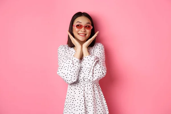 Concepto de turismo y estilo de vida. hermosa asiático mujer mostrando su limpio lindo cara, usando gafas de sol, sonriendo y mirando a un lado con admiración, de pie sobre rosa fondo — Foto de Stock